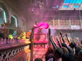 A vibrant celebration scene with a group of people gathered in a richly decorated setting. One person, elevated on a podium, is throwing pink colored powder into the air, while the crowd below reaches up towards it. There are decorations and lights, and the architecture is ornate with intricate designs and a chandelier hanging from the ceiling.