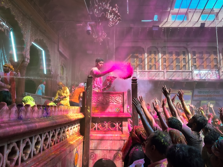 A vibrant celebration scene with a group of people gathered in a richly decorated setting. One person, elevated on a podium, is throwing pink colored powder into the air, while the crowd below reaches up towards it. There are decorations and lights, and the architecture is ornate with intricate designs and a chandelier hanging from the ceiling.