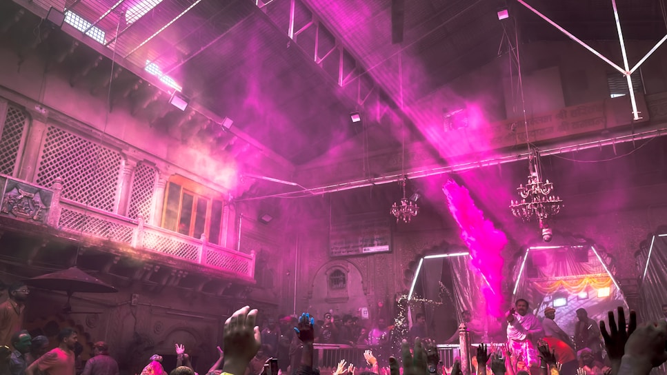 An indoor festival or celebration with people throwing vibrant pink and purple powder into the air. The area is packed with participants, and the scene is filled with colorful decorations. The setting has intricate architecture with balconies and chandeliers, giving it an old-world charm. The crowd is enthusiastic, with hands raised in joy and excitement.