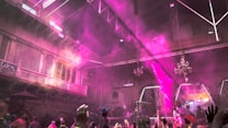 An indoor festival or celebration with people throwing vibrant pink and purple powder into the air. The area is packed with participants, and the scene is filled with colorful decorations. The setting has intricate architecture with balconies and chandeliers, giving it an old-world charm. The crowd is enthusiastic, with hands raised in joy and excitement.