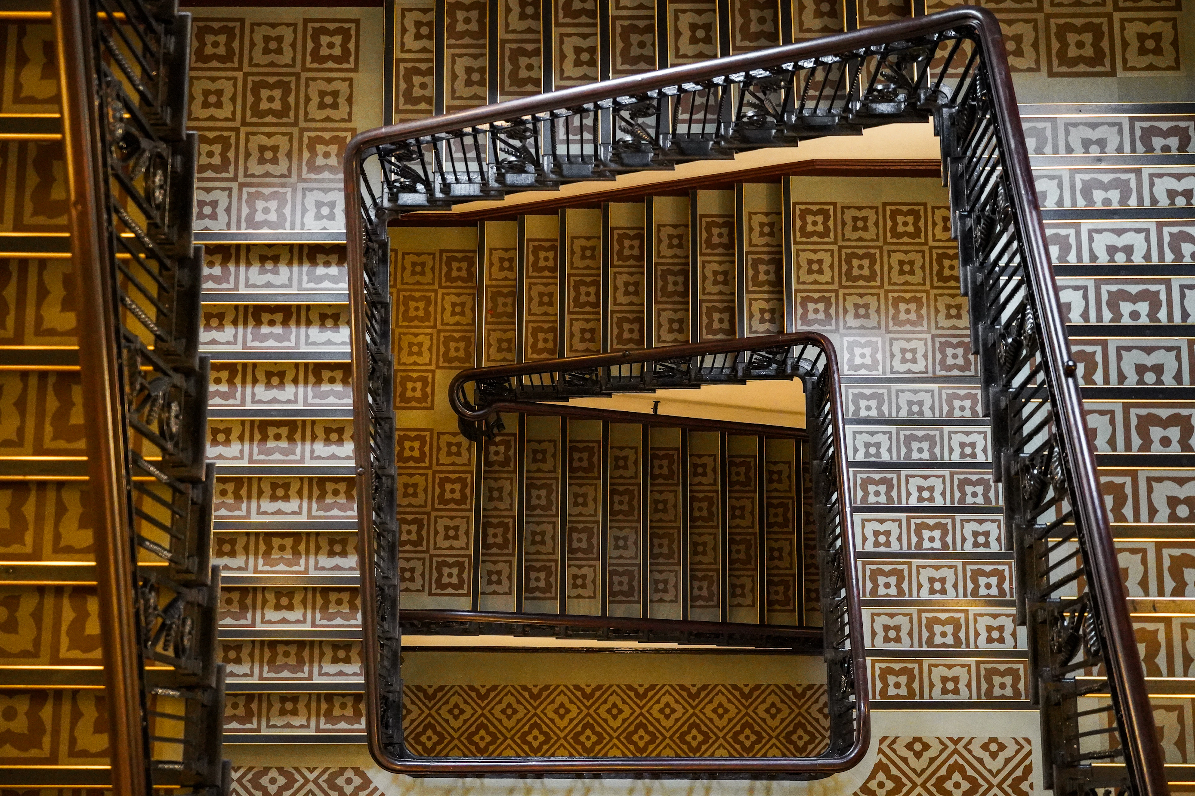 Looking down the square spiral staircase of the Titanic Hotel Belfast, dark polished handrails winding around a well lined with ochre and cream patterned tilework.