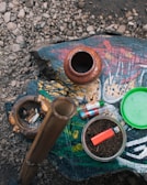 A rustic arrangement features various items on a painted wooden surface. An earthen pot and a carved ashtray with cigarette butts sit to the left. In the center, a bamboo pipe stands upright, and a glass bowl filled with tobacco and a red lighter are visible. Several colorful lighters lie nearby, with a green lid placed off to the side. The rough texture of gravel surrounds the main setup.