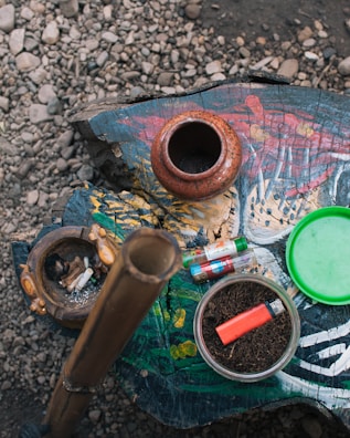 A rustic arrangement features various items on a painted wooden surface. An earthen pot and a carved ashtray with cigarette butts sit to the left. In the center, a bamboo pipe stands upright, and a glass bowl filled with tobacco and a red lighter are visible. Several colorful lighters lie nearby, with a green lid placed off to the side. The rough texture of gravel surrounds the main setup.