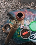 A rustic arrangement features various items on a painted wooden surface. An earthen pot and a carved ashtray with cigarette butts sit to the left. In the center, a bamboo pipe stands upright, and a glass bowl filled with tobacco and a red lighter are visible. Several colorful lighters lie nearby, with a green lid placed off to the side. The rough texture of gravel surrounds the main setup.