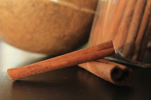 Close-up of several cinnamon sticks against a blurred background that includes a textured, rounded object and a plastic bag containing more cinnamon sticks.