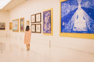 A person in a light pink dress stands in a spacious, well-lit art gallery observing a series of colorful framed paintings on the wall. The room features several artworks, including a prominent blue and gold painting depicting a serene, meditative figure. The gallery has a minimalist design with polished, reflective flooring.