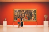 A man and a woman stand in an art gallery, admiring a large, colorful painting on a vibrant red wall. The painting is framed in an ornate golden frame and depicts an elaborate scene with several figures in classical attire. To the sides of the painting are two sculptures on white pedestals. The gallery is well-lit, with a modern and clean interior.