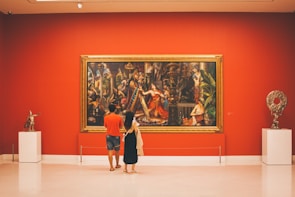 A man and a woman stand in an art gallery, admiring a large, colorful painting on a vibrant red wall. The painting is framed in an ornate golden frame and depicts an elaborate scene with several figures in classical attire. To the sides of the painting are two sculptures on white pedestals. The gallery is well-lit, with a modern and clean interior.