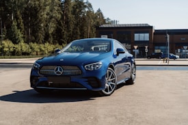 A sleek, blue luxury car parked on a sunlit concrete surface, surrounded by trees and a modern building in the background. The car's stylish design and logo are prominently displayed.