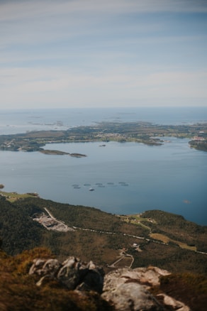 A vast and serene landscape with a clear view of a coastal area. The image features calm blue waters with several circular fish farm installations dotting the surface. In the foreground, there is rugged terrain with patches of greenery. The background consists of distant islands and landmasses under a lightly clouded sky.