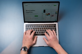 A pair of hands are typing on a laptop keyboard, with a website displayed on the screen. The background is a solid blue surface. The left hand is wearing a wristwatch, and the laptop is open and actively being used.