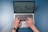A pair of hands are typing on a laptop keyboard, with a website displayed on the screen. The background is a solid blue surface. The left hand is wearing a wristwatch, and the laptop is open and actively being used.