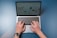 A pair of hands are typing on a laptop keyboard, with a website displayed on the screen. The background is a solid blue surface. The left hand is wearing a wristwatch, and the laptop is open and actively being used.