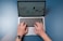 A pair of hands are typing on a laptop keyboard, with a website displayed on the screen. The background is a solid blue surface. The left hand is wearing a wristwatch, and the laptop is open and actively being used.