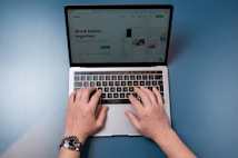 A pair of hands are typing on a laptop keyboard, with a website displayed on the screen. The background is a solid blue surface. The left hand is wearing a wristwatch, and the laptop is open and actively being used.