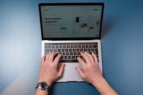 A pair of hands are typing on a laptop keyboard, with a website displayed on the screen. The background is a solid blue surface. The left hand is wearing a wristwatch, and the laptop is open and actively being used.