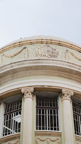 A historic building displays classical architectural elements, featuring ornate columns with Corinthian capitals and decorative reliefs. Cracks and peeling paint indicate weathering and age, adding character to the structure. Two windows with detailed iron grilles stand out against the faded, light-colored facade.