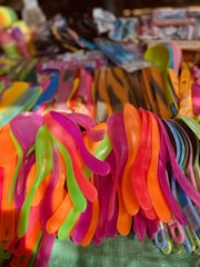 A collection of colorful plastic utensils stacked together, including neon-colored spoons and spatulas. The vibrant hues of pink, orange, green, and purple create a lively and cheerful atmosphere. The utensils are arranged in a crowded display, suggesting a market or shop setting.