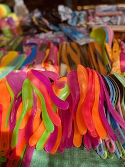 A collection of colorful plastic utensils stacked together, including neon-colored spoons and spatulas. The vibrant hues of pink, orange, green, and purple create a lively and cheerful atmosphere. The utensils are arranged in a crowded display, suggesting a market or shop setting.