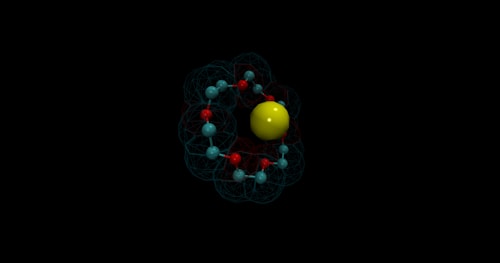 A 3D representation of an atom structure featuring a central yellow sphere surrounded by smaller light blue spheres connected via red rods. The spheres and rods form a network resembling electron paths or orbitals, all set against a dark background.