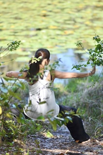 A person wearing a white tank top and dark pants is stretching or practicing a form of exercise outdoors, surrounded by green foliage and a pond with lily pads in the background. Their hair is tied back in a ponytail, and they appear to be focused and calm, engaging with nature.