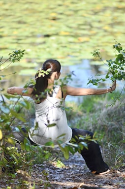 A person wearing a white tank top and dark pants is stretching or practicing a form of exercise outdoors, surrounded by green foliage and a pond with lily pads in the background. Their hair is tied back in a ponytail, and they appear to be focused and calm, engaging with nature.