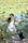 A person wearing a white tank top and dark pants is stretching or practicing a form of exercise outdoors, surrounded by green foliage and a pond with lily pads in the background. Their hair is tied back in a ponytail, and they appear to be focused and calm, engaging with nature.