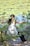 A person wearing a white tank top and dark pants is stretching or practicing a form of exercise outdoors, surrounded by green foliage and a pond with lily pads in the background. Their hair is tied back in a ponytail, and they appear to be focused and calm, engaging with nature.