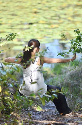 A person wearing a white tank top and dark pants is stretching or practicing a form of exercise outdoors, surrounded by green foliage and a pond with lily pads in the background. Their hair is tied back in a ponytail, and they appear to be focused and calm, engaging with nature.