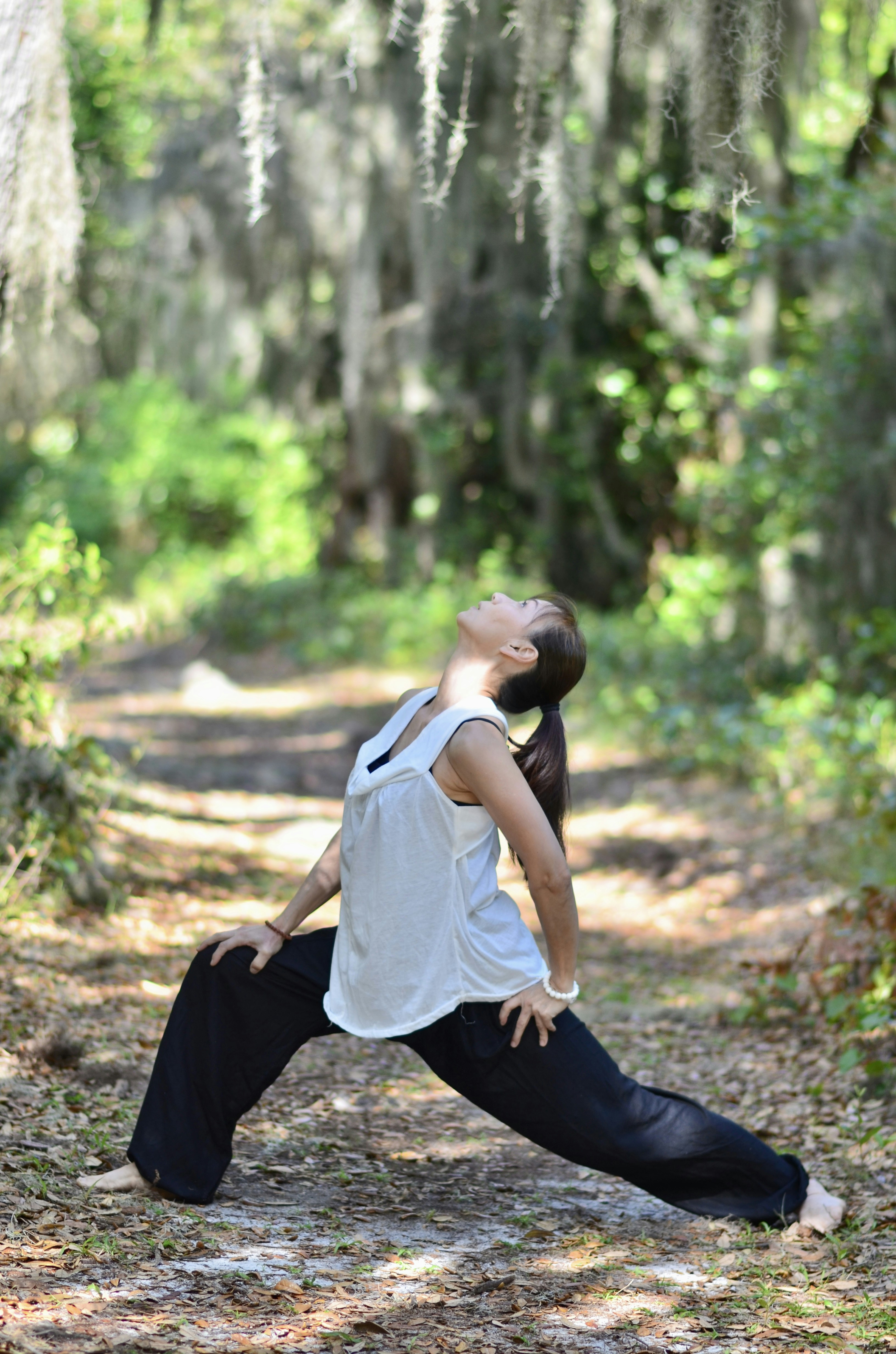 A Japanese American person with a long ponytail, bracelets, black loose pants and a white tank top does qigong in a forest.