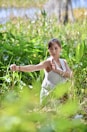 A person is practicing a martial art form in a lush green natural setting. Surrounded by tall grass and foliage, they hold a poised stance with focused concentration. Sunlight filters through the leaves, creating a serene and tranquil atmosphere.