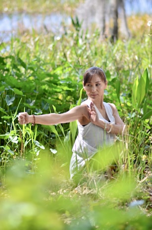 A person is practicing a martial art form in a lush green natural setting. Surrounded by tall grass and foliage, they hold a poised stance with focused concentration. Sunlight filters through the leaves, creating a serene and tranquil atmosphere.