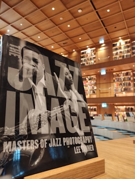 A large book titled 'The Jazz Image' with a black and white photograph on the cover stands on a wooden holder. The background reveals a spacious, warmly lit library with tall bookshelves and a modern interior design.