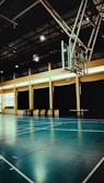 An indoor sports hall with a basketball hoop and court markings. The court has a polished wooden floor with red and white lines. The ceiling is high with metal beams, and the walls have large windows covered by dark curtains. A basketball is visible in mid-air near the hoop.
