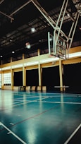 An indoor sports hall with a basketball hoop and court markings. The court has a polished wooden floor with red and white lines. The ceiling is high with metal beams, and the walls have large windows covered by dark curtains. A basketball is visible in mid-air near the hoop.