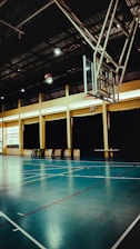An indoor sports hall with a basketball hoop and court markings. The court has a polished wooden floor with red and white lines. The ceiling is high with metal beams, and the walls have large windows covered by dark curtains. A basketball is visible in mid-air near the hoop.