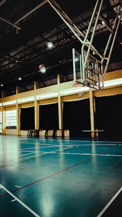 An indoor sports hall with a basketball hoop and court markings. The court has a polished wooden floor with red and white lines. The ceiling is high with metal beams, and the walls have large windows covered by dark curtains. A basketball is visible in mid-air near the hoop.
