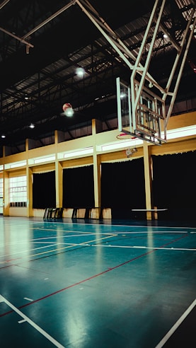 An indoor sports hall with a basketball hoop and court markings. The court has a polished wooden floor with red and white lines. The ceiling is high with metal beams, and the walls have large windows covered by dark curtains. A basketball is visible in mid-air near the hoop.