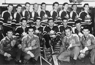 A vintage black and white photograph featuring a group of men in ice hockey uniforms and jackets, arranged in three rows. The men are part of a sports team, with those in the back two rows wearing jerseys with a maple leaf emblem and the word 'Schrider' printed on them. The front row includes individuals in jackets with similar emblems sitting cross-legged on the floor. An ice hockey stick is visible at the bottom, and the backdrop appears to be in a locker room or team area.