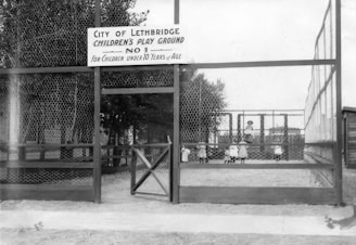 A fenced playground with a sign indicating it is for children under 10 years of age. The play area features several children in early 20th-century clothing, engaging on a swing set. The structure and surrounding fence are made of wood and wire mesh, with trees visible in the background.