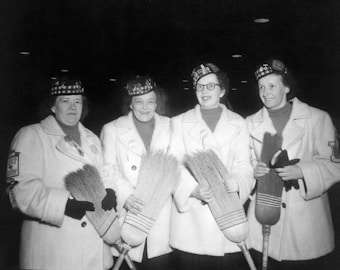 Four women are standing together, each holding a curling broom. They are wearing matching coats and plaid hats, suggesting a team or uniform look. The setting appears to be indoors with a dark background and overhead lights, indicating a sports or recreational event.