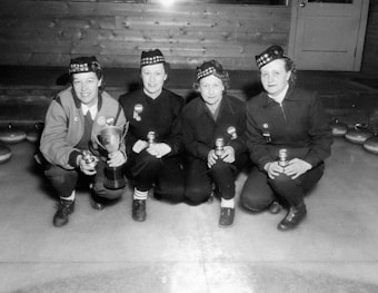 Four individuals are crouching on a curling rink, dressed in matching uniforms and plaid hats. One person is holding a trophy, while the others hold soda cans. Curling stones are visible behind them, and a wooden wall and door are in the background.