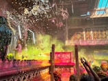 A vibrant scene filled with colored powders and splashes of water. People are participating in what seems to be a festive celebration inside a traditional architectural setting adorned with intricate carvings and grand chandeliers. The atmosphere is lively and crowded with participants throwing colors and raising their hands in joy.