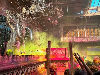 A vibrant scene filled with colored powders and splashes of water. People are participating in what seems to be a festive celebration inside a traditional architectural setting adorned with intricate carvings and grand chandeliers. The atmosphere is lively and crowded with participants throwing colors and raising their hands in joy.