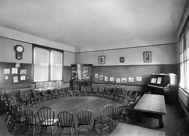 A vintage classroom setup with a large circular arrangement of wooden chairs around an empty space, suggesting a gathering or discussion area. The room features large windows with blinds, letting in natural light. A wall displays various artworks and photographs, adding an educational atmosphere. A piano with sheet music sets to the side, and a clock is mounted on the wall.