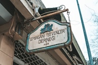 An aged metal sign with a blue outline hangs from chains, displaying the text 'EMİRGAN'I SEVENLER DERNEĞİ 2005' and a leaf emblem above it. The setting appears to be outdoors, with a building structure and some electrical wires visible, suggesting an urban environment.