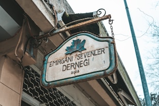 An aged metal sign with a blue outline hangs from chains, displaying the text 'EMİRGAN'I SEVENLER DERNEĞİ 2005' and a leaf emblem above it. The setting appears to be outdoors, with a building structure and some electrical wires visible, suggesting an urban environment.
