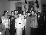 A group of elegantly dressed women stand together in a formal setting. They are wearing traditional Asian attire with intricate designs and patterns. In the background, two men in suits are visible among various flags and a portrait on the wall. The setting appears to be indoors, possibly at a cultural or diplomatic event.
