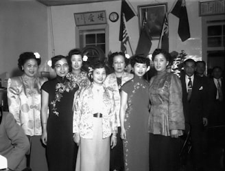 A group of elegantly dressed women stand together in a formal setting. They are wearing traditional Asian attire with intricate designs and patterns. In the background, two men in suits are visible among various flags and a portrait on the wall. The setting appears to be indoors, possibly at a cultural or diplomatic event.