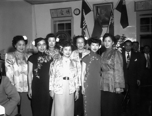 A group of elegantly dressed women stand together in a formal setting. They are wearing traditional Asian attire with intricate designs and patterns. In the background, two men in suits are visible among various flags and a portrait on the wall. The setting appears to be indoors, possibly at a cultural or diplomatic event.